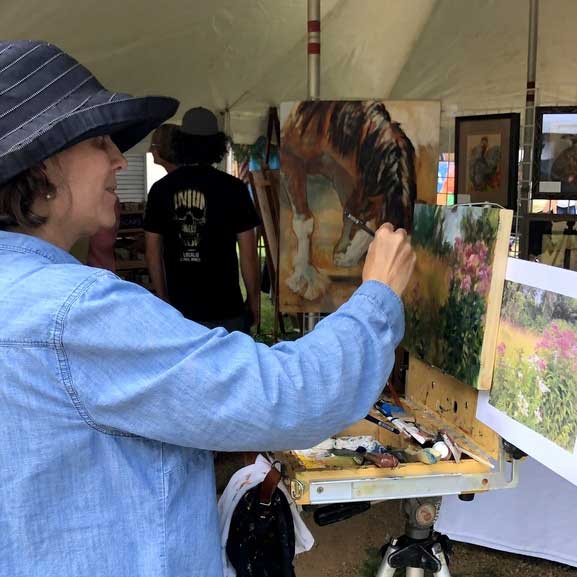 A woman wearing a hat stands at a paint easel and paints a nature scene using a photograph image as the source of inspiration.