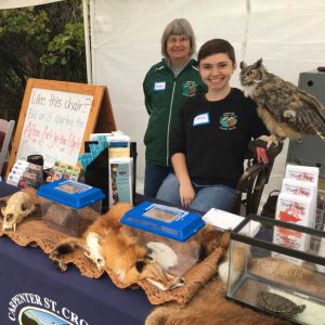 Two women are behind an educational display table with live animals including a great-horned owl, a turtle, and a reptile. There are other nature-themed items on display.