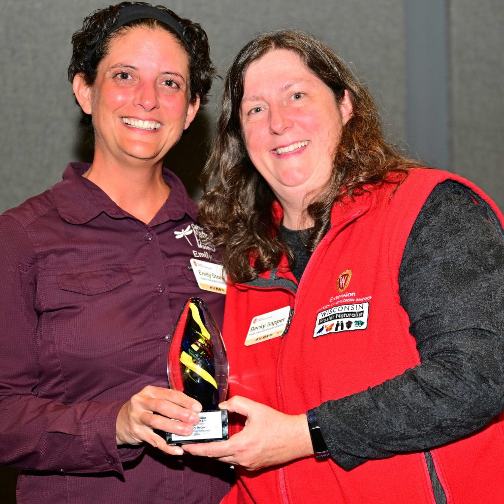 two women smile at the camera during an award ceremony. The woman on the right wears a red vest with a Master Naturalist badge. The woman on the left is wearing a burgundy shirt with the Cable Natural History Museum logo on the chest.