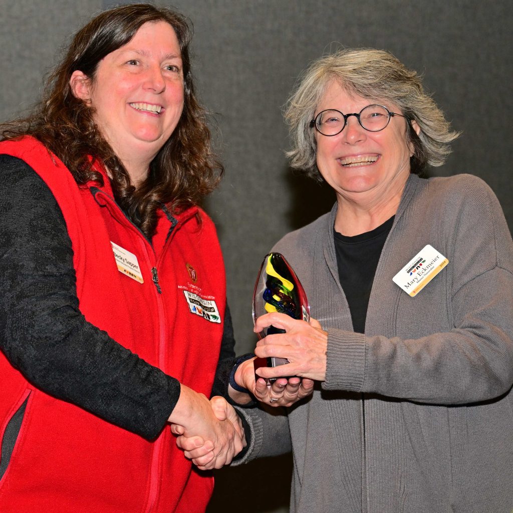 Two women smile during an award ceremony. The woman on the left is wearing a red vest with the Master Naturalist logo. The woman on the right wears glasses and a grey sweater as has the biggest smile as she receives the award.