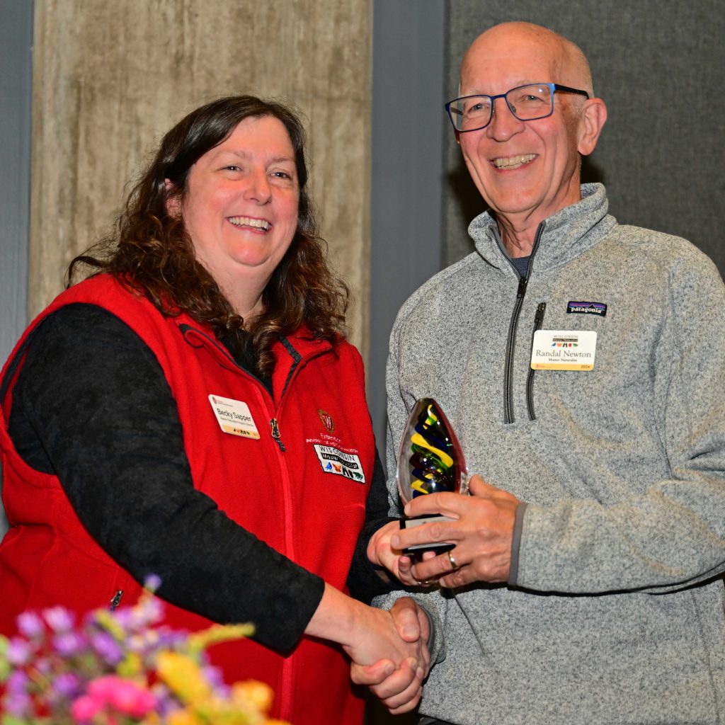 A woman and a man smile for the camera during an awards ceremony. The woman is wearing a red vest with the Master Naturalist logo and shakes the man's hand as he receives the award from her.
